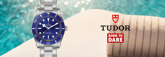 A Tudor wristwatch with a blue dial and blue bezel, shown beside a rolled beige towel with the Tudor logo and 'Born to Dare' text against a pool-water background.